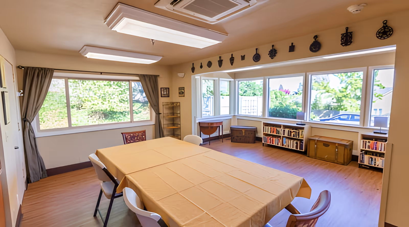 Bright activity/dining room with a large table covered by a beige tablecloth, chairs, bookshelves under wide windows, and decorative wall accents.