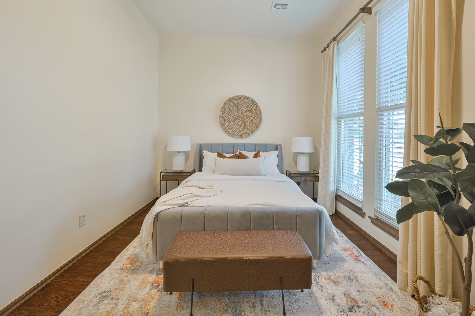 Neatly made bedroom with a bed centered between two nightstands, large windows with blinds and curtains, and a decorative plant.