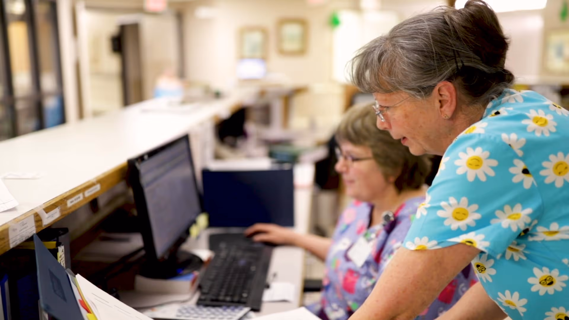 Two healthcare workers in colorful scrubs working together at a computer station in a medical or senior care facility. One is seated typing on the keyboard while the other leans over to assist or discuss something on the screen.