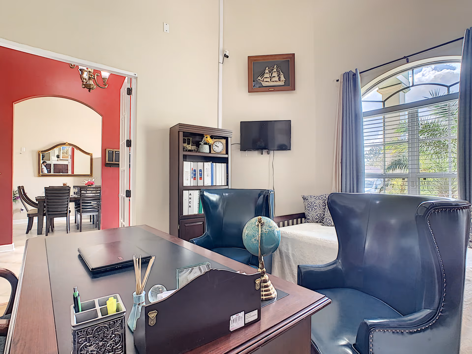An office area in a senior living facility with a wooden desk holding a laptop, globe, and office supplies. Two blue leather wingback chairs are positioned near the desk. A small bookshelf with binders and decorative items is against the wall. A flat-screen TV is mounted on the wall above a daybed with patterned pillows. A large window with blue curtains lets in natural light, and an adjacent room with a dining table and chairs is visible through an open doorway with red walls.