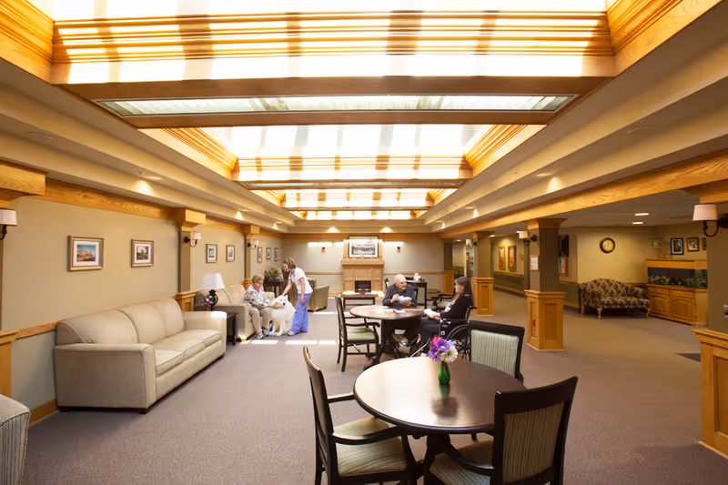 A bright and spacious common area in a senior living facility with a high ceiling featuring skylights. The room has beige walls, wooden trim, and carpeted floors. There are several round tables with chairs, a beige sofa, and a fireplace at the far end. Two elderly people are seated at a table, one in a wheelchair, while a caregiver interacts with another elderly person sitting on a chair with a white dog. The room is decorated with framed pictures on the walls and a vase with flowers on one of the tables.