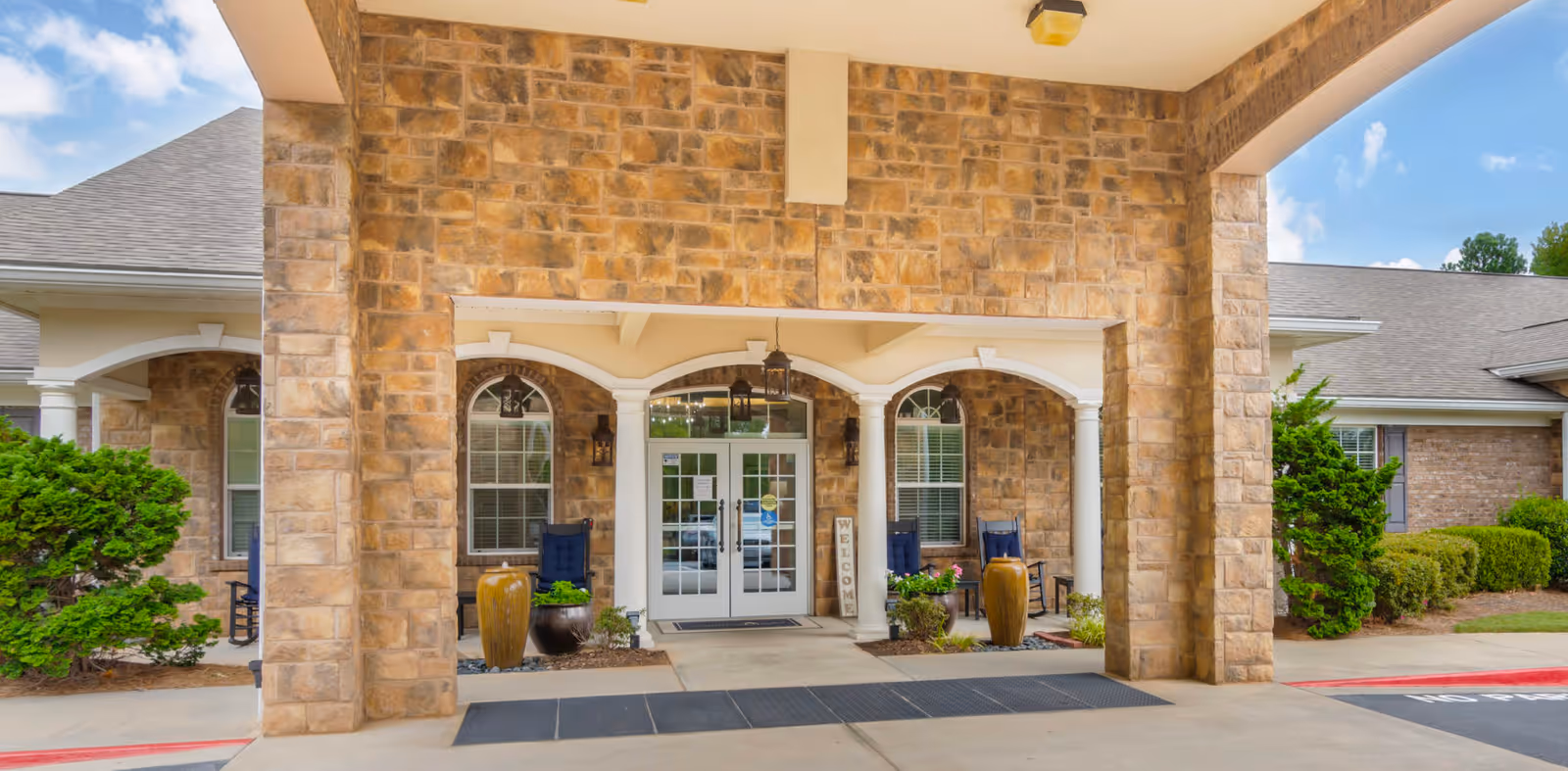 Covered stone entrance to a senior living facility showing double glass doors, arched windows, potted plants, and seating.