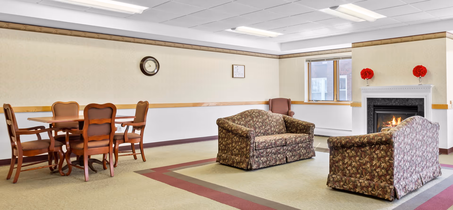 A senior living facility common area with two floral-patterned sofas facing a lit fireplace. There is a wooden table with four chairs on the left side, a clock on the wall, a window letting in natural light, and two red floral decorations on the fireplace mantel.