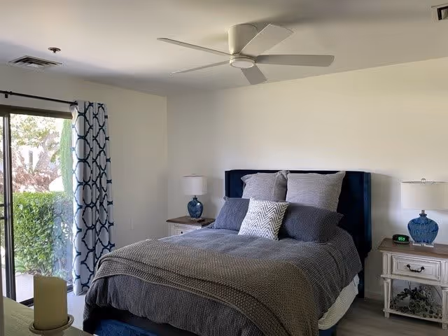 A neatly made bed with gray bedding and multiple pillows in a bedroom. There are two bedside tables with blue lamps and an alarm clock on the right table. A ceiling fan is mounted above the bed, and a window with patterned curtains shows greenery outside.