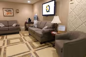 Comfortable seating area with two sofas, an armchair, a wall-mounted TV, lamp, and side tables in a nursing facility lounge.