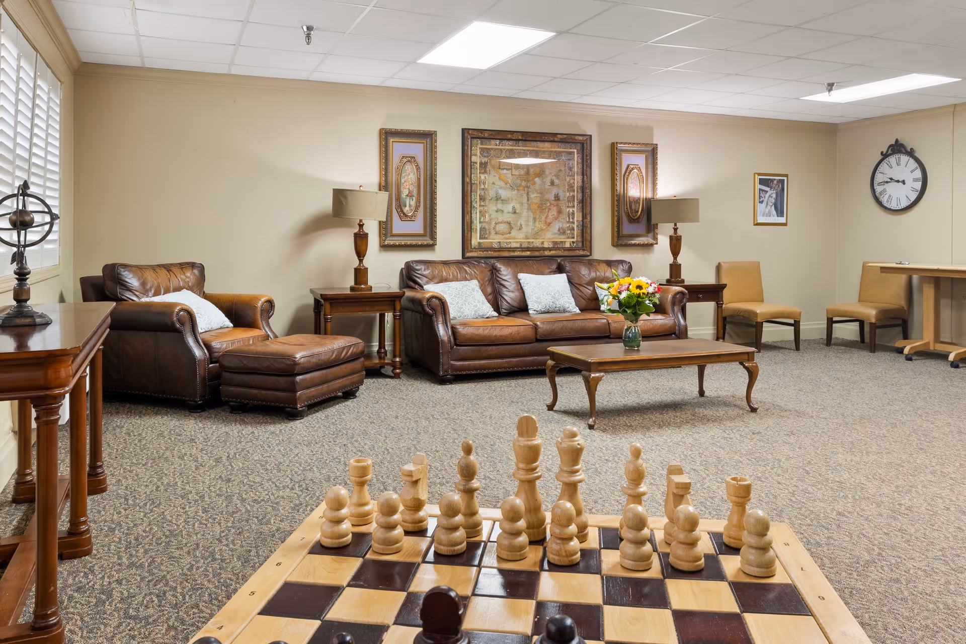 A cozy living room with brown leather sofa and armchair, a wooden coffee table with a vase of flowers, two side tables with lamps, framed artwork on the wall, a large chessboard in the foreground, and additional chairs and a clock on the far wall.