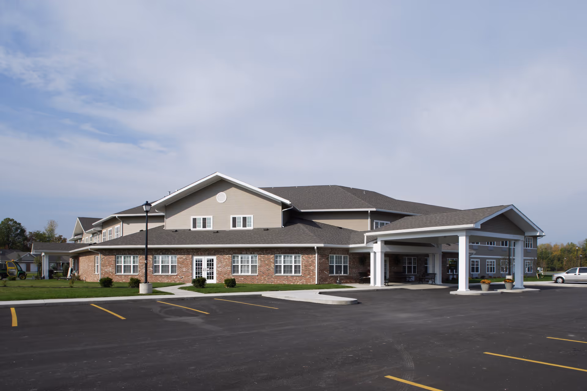 Exterior view of Elderwood Residences at Wheatfield, a two-story senior living facility with a brick and beige siding facade, a covered entrance, and a parking lot in front under a partly cloudy sky.