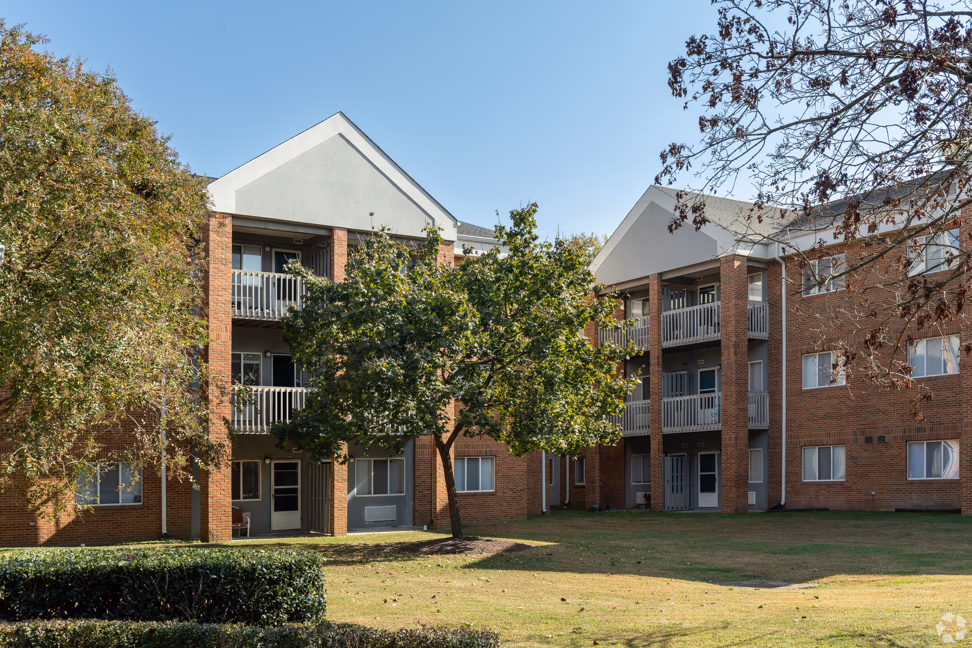 Exterior view of a three-story brick residential building with balconies and multiple windows, surrounded by trees and a grassy lawn under a clear blue sky.