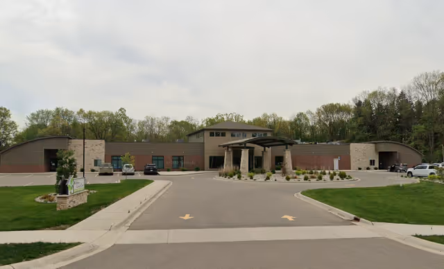 Front exterior of Serene Gardens of Clarkston showing a single-story brick building with a covered entrance, circular drive, parked cars, and landscaped grounds.