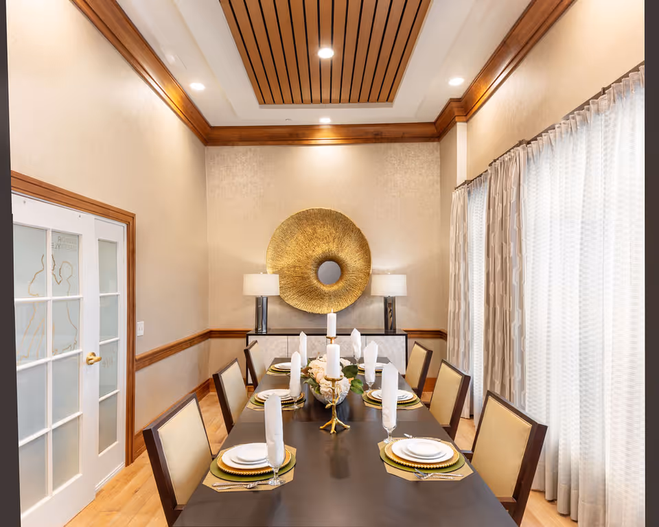 A formal dining room with a long dark wooden table set for eight people with white plates, gold placemats, folded white napkins, and glassware. The room features beige walls with wooden trim, a decorative gold circular wall piece, two table lamps on a sideboard, and large windows with patterned curtains allowing natural light.