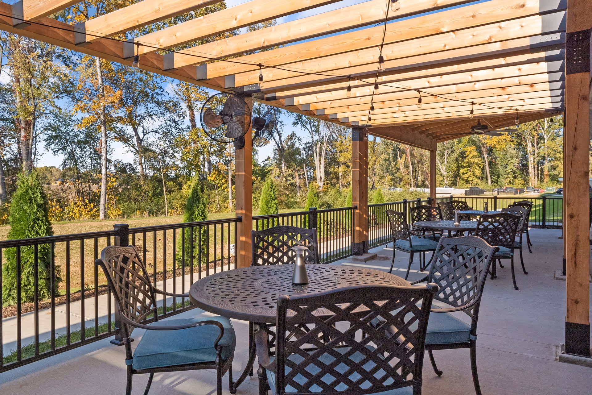 Covered outdoor patio with metal dining tables and chairs under a wooden pergola overlooking a tree-lined lawn.