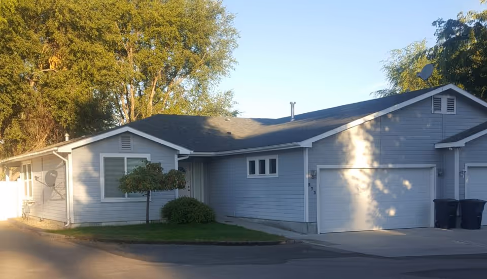 Exterior view of a single-story residential building with light blue siding, a dark shingled roof, a garage with a white door, and a small tree and shrub in front. The building is surrounded by trees and is illuminated by sunlight.