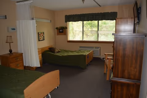 A nursing facility room with two single beds covered in green bedspreads, a wooden nightstand with a lamp, a wooden dresser, a chair, a large window with green valance curtains, and a privacy curtain partially drawn between the beds.