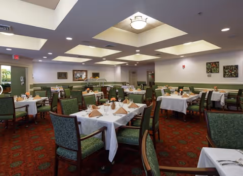 A spacious dining room with multiple tables covered in white tablecloths, each set with folded beige napkins, glasses, and small floral centerpieces. The room has green cushioned chairs, a red patterned carpet, and a ceiling with recessed lighting and a central light fixture. There are framed pictures on the walls and a door leading outside.
