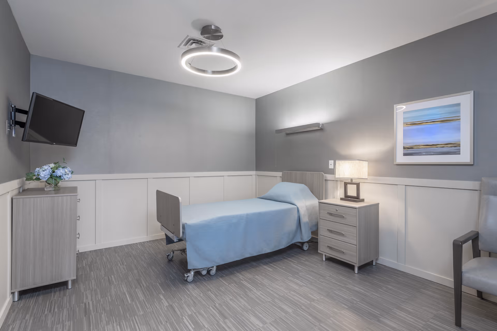 A simple, clean bedroom in a senior living facility featuring a single bed with light blue bedding, a bedside table with a lamp, a wall-mounted TV, a small dresser with a vase of flowers, a gray armchair, and a framed abstract painting on the wall. The room has gray walls with white wainscoting and a modern circular ceiling light.