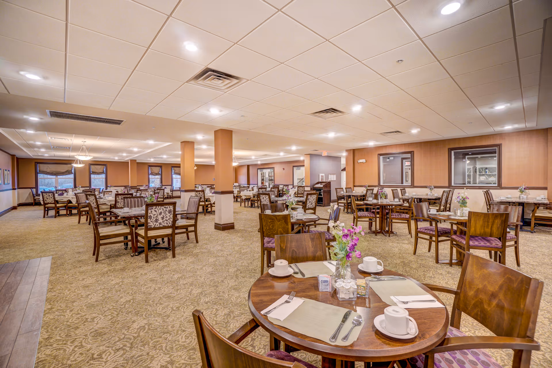A spacious dining room in a senior living facility with multiple round and rectangular tables set with cups, saucers, utensils, and floral centerpieces. The room has carpeted floors, beige walls, and a drop ceiling with recessed lighting. Several windows and columns are visible throughout the room.