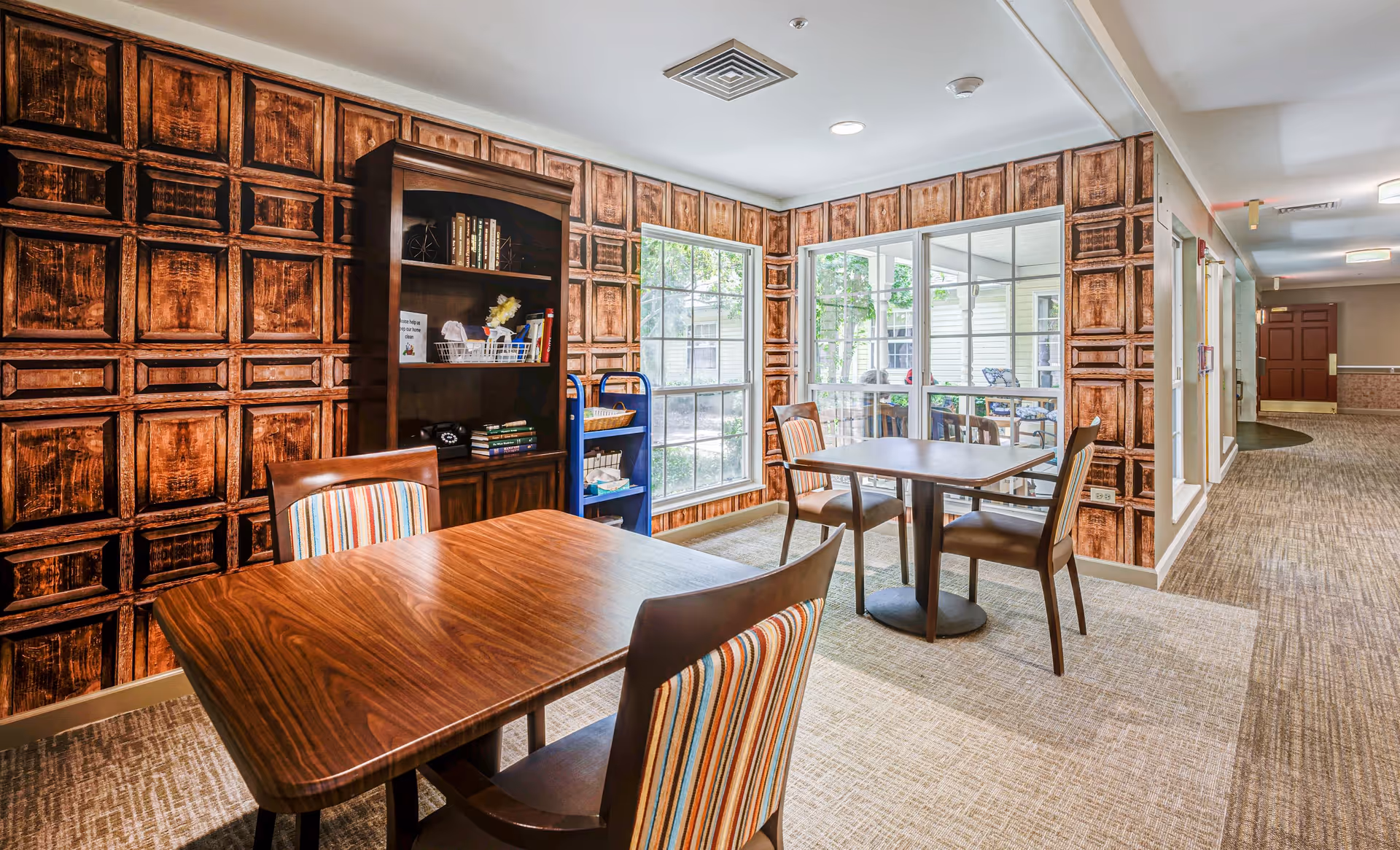 Interior view of a senior living facility room with two wooden tables and four chairs with striped upholstery. The walls have a decorative wood panel pattern, and there is a bookshelf with books and decorative items. Large windows let in natural light and show an outdoor patio area with seating. The room has carpeted floors and a hallway leading to other parts of the facility.