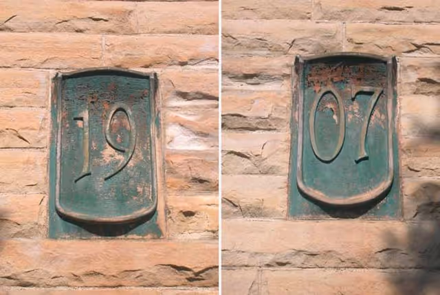 Two weathered green metal plaques set into a stone wall displaying the numbers '19' and '07'.