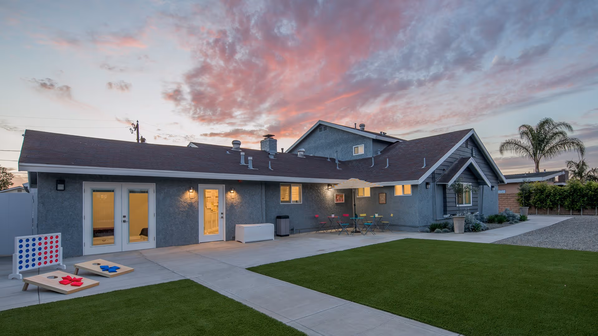 Outdoor backyard and patio of a single-story residential care building at sunset, with artificial turf, patio games, and seating.