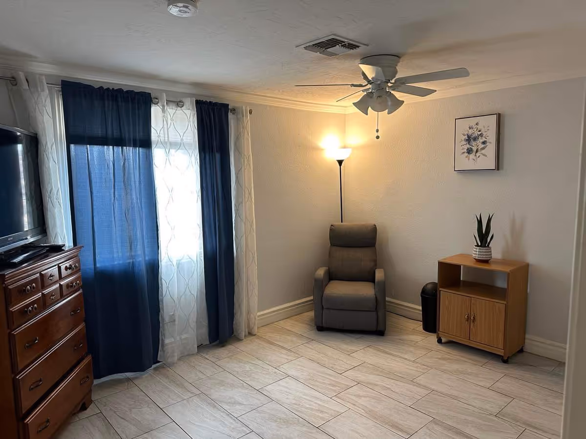 A cozy room with light-colored tiled flooring and light gray walls. There is a gray recliner chair in the corner next to a tall floor lamp emitting warm light. A small wooden cabinet with a potted plant on top is placed against the wall. A framed floral artwork hangs above the cabinet. On the left side, there is a wooden dresser with a flat-screen TV on top, and a window covered with dark blue and sheer white curtains.