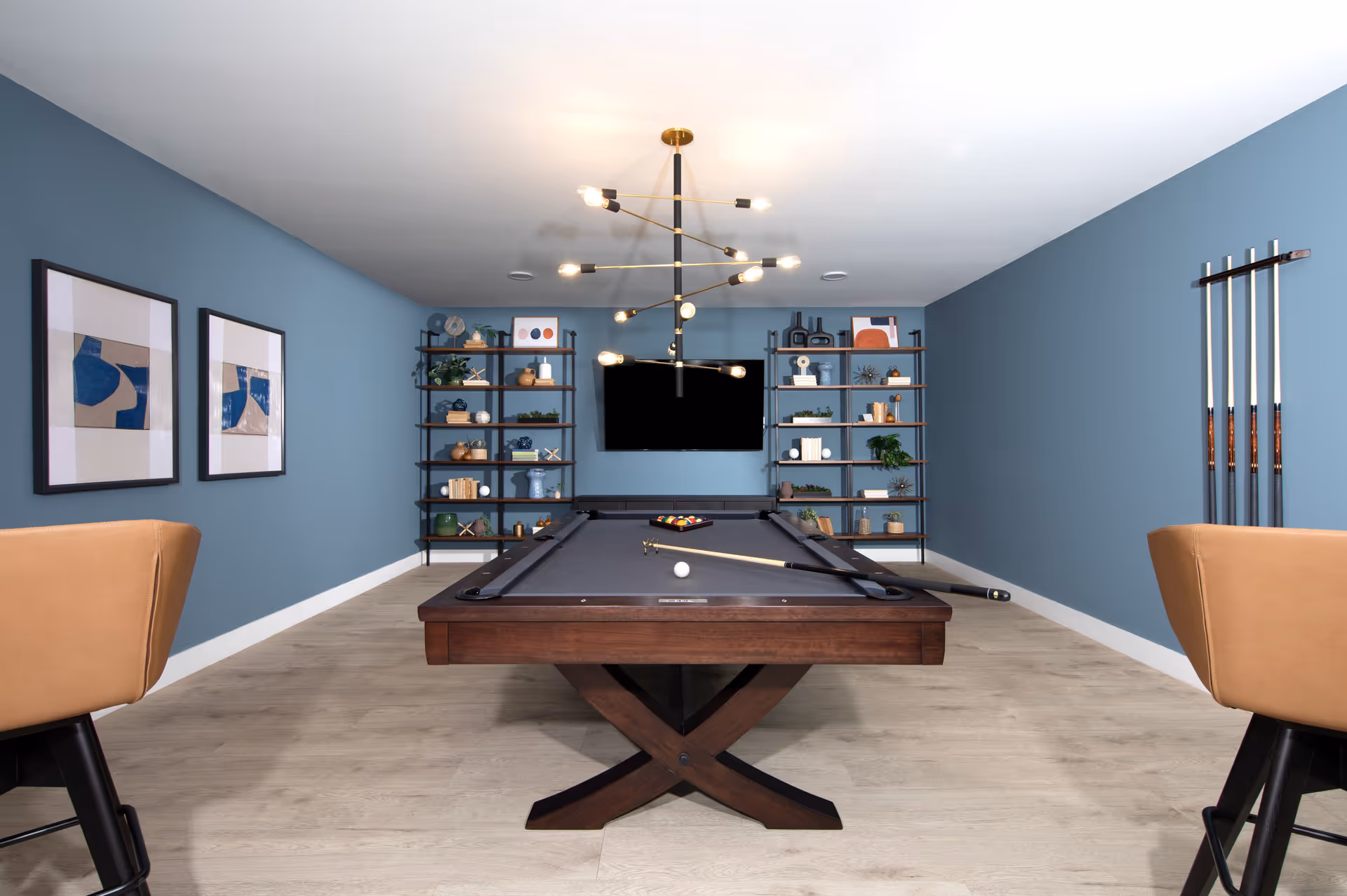 A modern game room with a pool table centered in the room. The pool table has a dark wood frame and a gray felt surface with a cue stick and a white ball on it. The walls are painted blue and decorated with two abstract framed artworks on the left and a rack holding pool cues on the right. At the far end, there are two black metal shelves filled with decorative items and a flat-screen TV mounted on the wall. Two tan leather chairs are partially visible on either side of the pool table. A contemporary chandelier with exposed bulbs hangs from the ceiling.