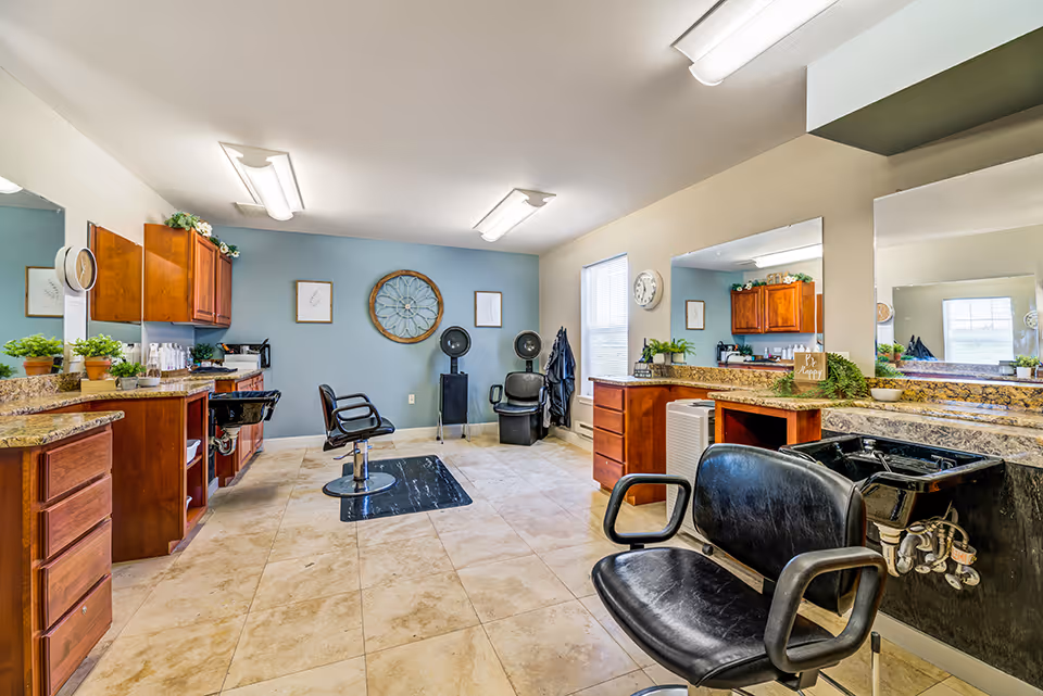 Interior view of a hair salon area in a senior living facility with salon chairs, sinks for washing hair, wooden cabinets, large mirrors, and decorative plants. The room has tiled floors, overhead fluorescent lighting, and a blue accent wall with framed artwork and a decorative circular wall piece.