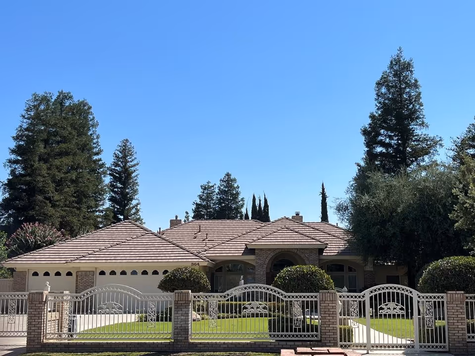 Single-story brick house with a tiled roof and decorative metal front gate enclosing a manicured lawn under a clear blue sky.