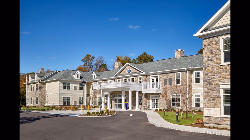 Exterior front view of Chelsea Senior Living at Washington Township, showing a large two-story building with stone and siding facade, multiple windows, a covered entrance with white columns, and a driveway with landscaping under a clear blue sky.