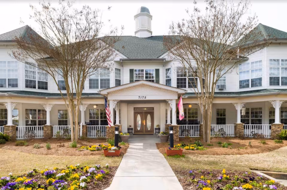Front exterior view of a two-story senior living facility with white siding, green roof, and a covered entrance. There are two leafless trees flanking the walkway leading to the double front doors. Flower beds with colorful flowers line the walkway and the front of the building. An American flag and another flag are displayed near the entrance.