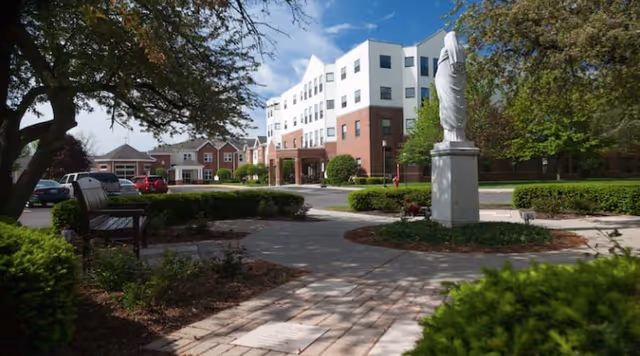 Paved courtyard with a statue, bench, and landscaping in front of a multi-story brick-and-white senior living building.