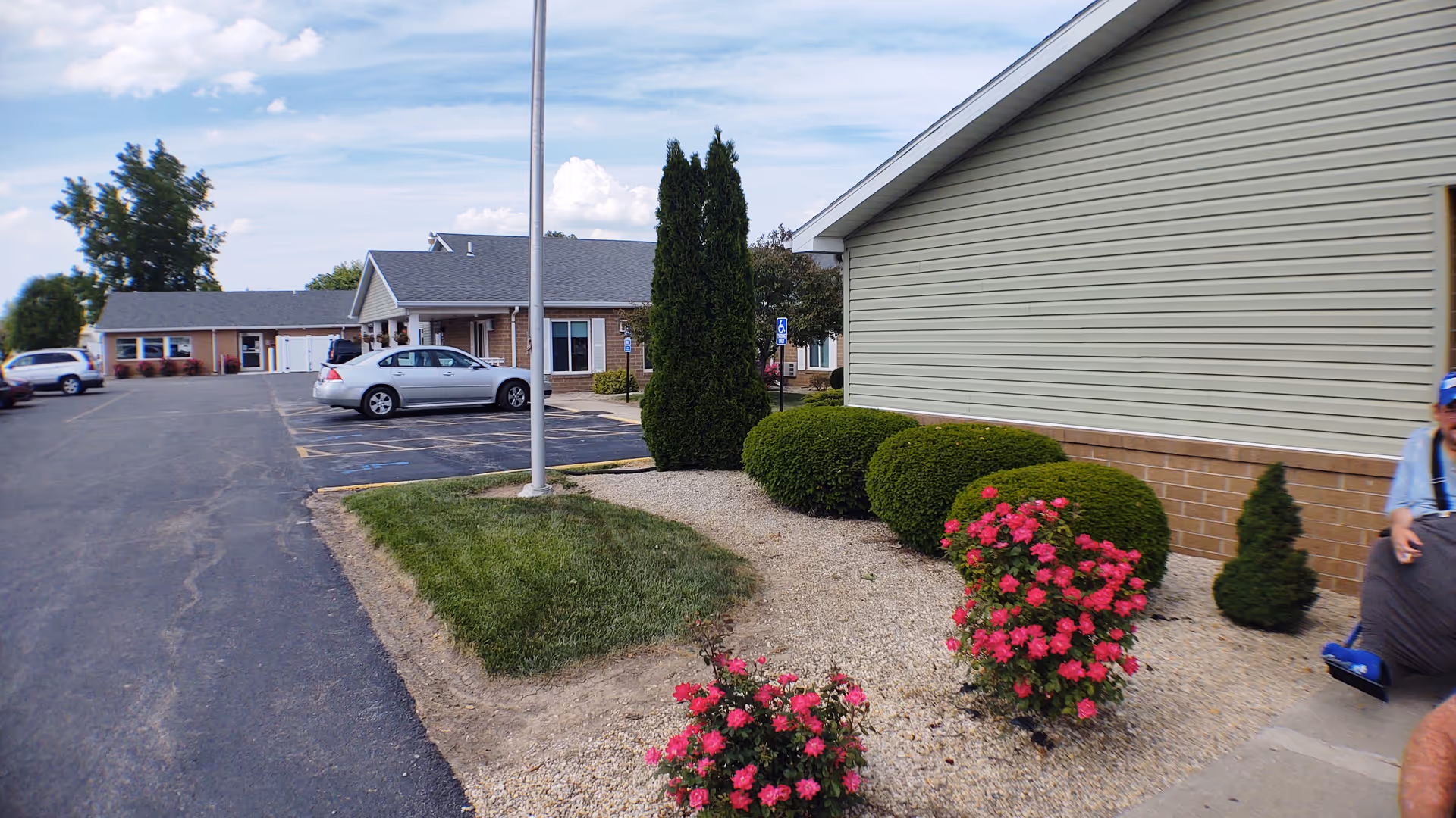 Exterior view of Windsor Lane Health Care facility showing a parking lot with several parked cars, a landscaped area with bushes and pink flowers, and part of the building with beige siding and brick base under a partly cloudy sky.
