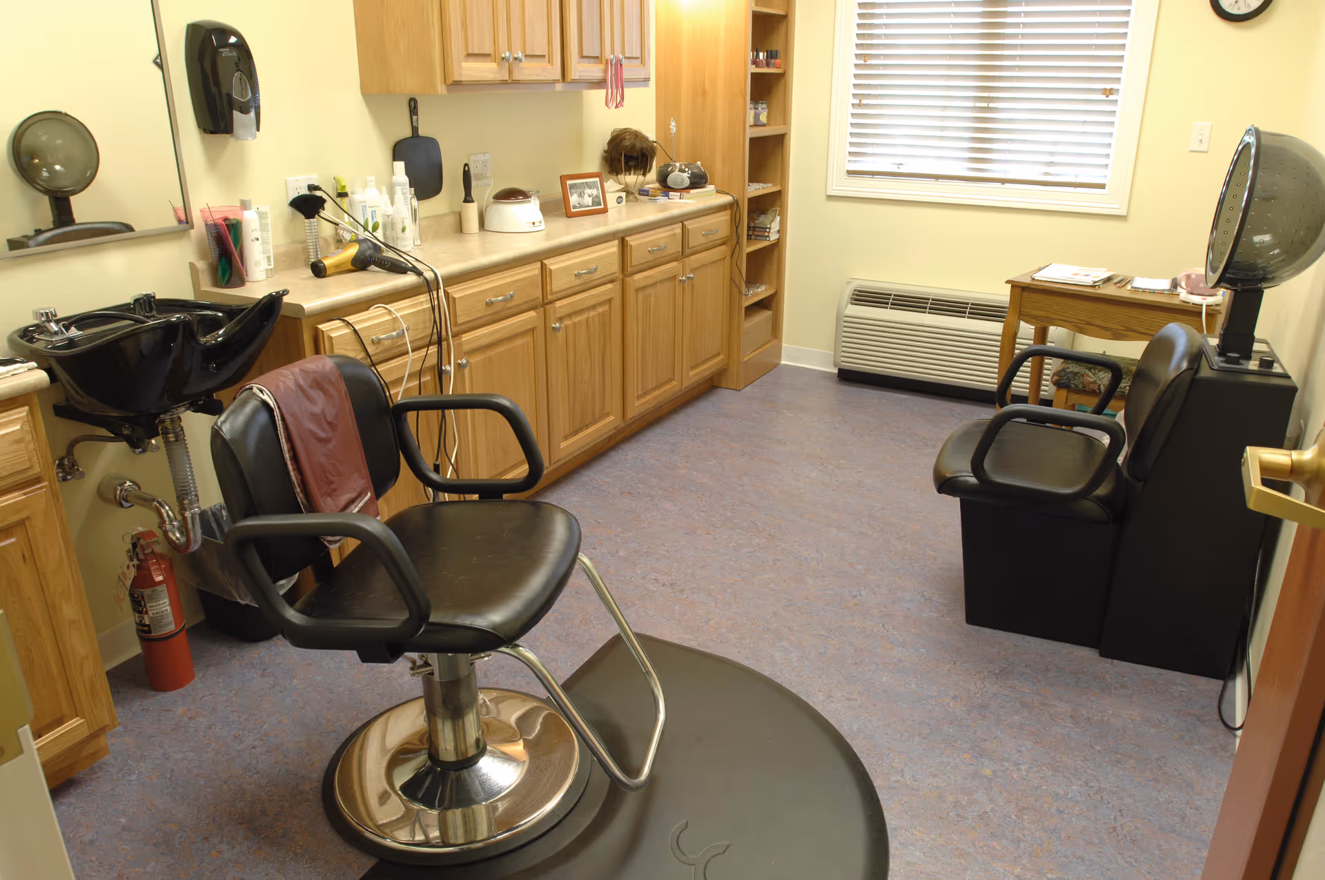 Interior view of a small hair salon or beauty room with a black salon chair in front of a black hair washing sink. The room has wooden cabinets along one wall with various hair care products, a hair dryer, and a wig on the counter. There is a window with blinds, a small wooden table with magazines, and another black salon chair with a hooded hair dryer.