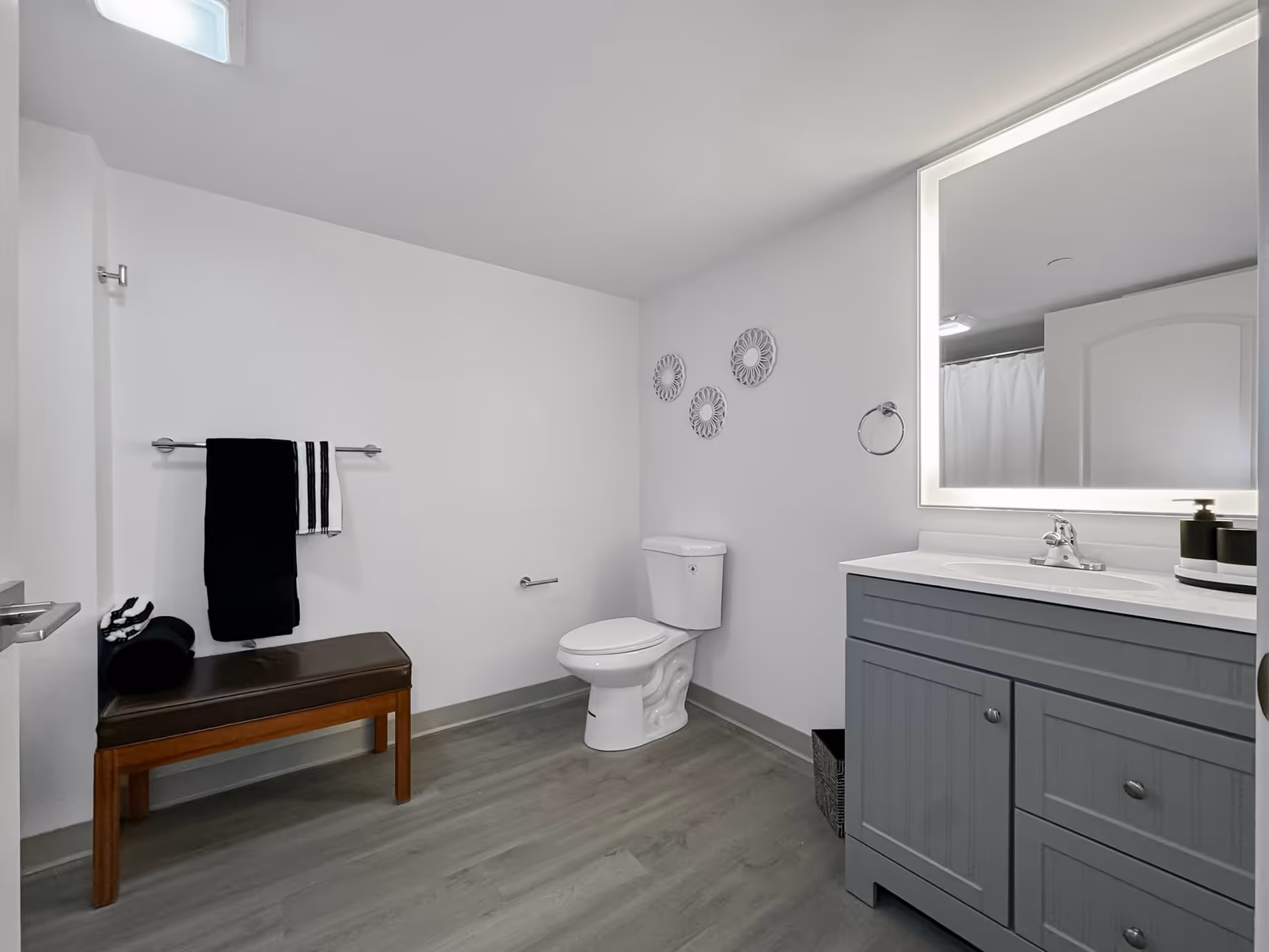 A modern bathroom with a white toilet, a gray vanity with a sink, and a large illuminated mirror above it. There is a wooden bench with a dark cushion and folded towels on it, and a towel rack with black and white towels hanging. The walls are white with three decorative circular wall hangings above the toilet. The floor is light gray wood.