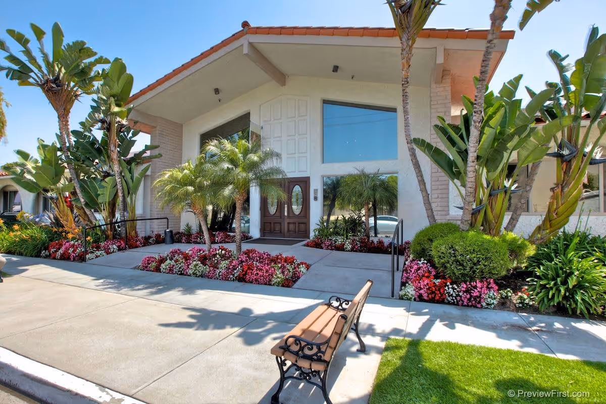 Front exterior view of Brittany House Residential Memory Care facility featuring a mid-century modern style building with large windows, a double wooden door entrance, surrounded by landscaped flower beds, palm trees, and a bench on the sidewalk.