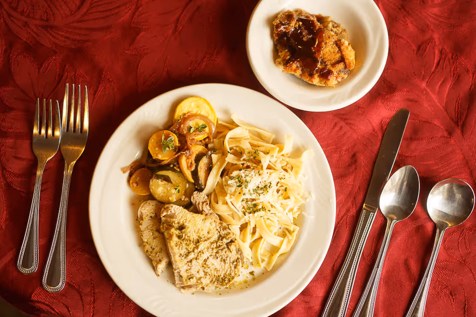 A white plate with a meal consisting of sliced turkey, cooked yellow squash and zucchini, and a serving of fettuccine pasta topped with herbs and cheese, placed on a red tablecloth. Next to the plate is a small white dish with a dessert topped with sauce. Silverware including two forks, a knife, and two spoons are arranged on either side of the plate.