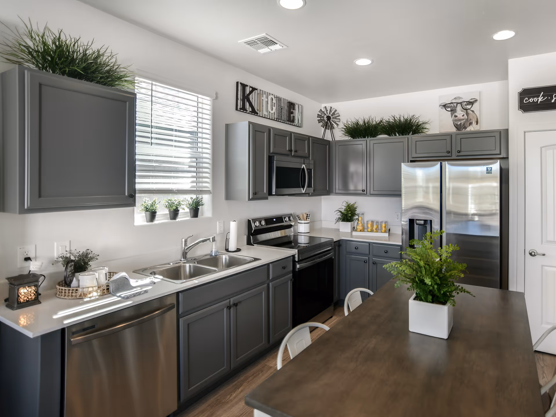 A modern kitchen with gray cabinets, stainless steel appliances including a refrigerator, oven, microwave, and dishwasher. There is a double sink under a window with small potted plants on the windowsill. The kitchen has a wooden island with a green potted plant on top and white chairs around it. Decorative items include a sign that spells 'KITCHEN', a small windmill, and a painting of a cow wearing glasses.