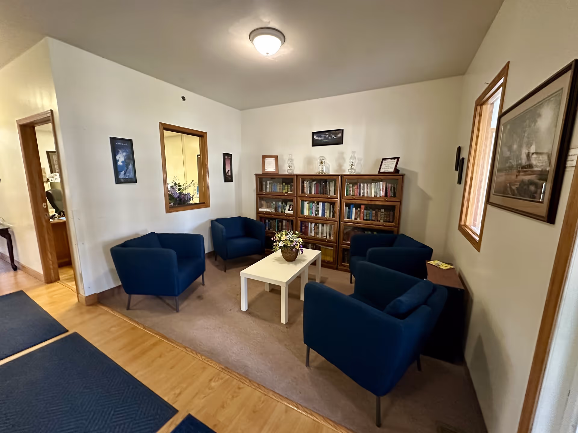 A cozy sitting area with four blue armchairs arranged around a white coffee table with a flower pot on it. Behind the chairs is a wooden bookshelf filled with books and decorative items. The room has light-colored walls with framed pictures and a window looking into another room. The floor is a combination of wood and carpet.