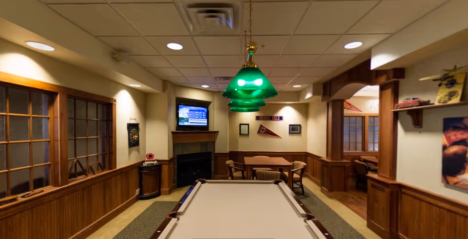 A cozy game room featuring a pool table in the center with green hanging lights above it. The room has wood paneling on the lower walls and beige wallpaper above. There is a small table with chairs in the back corner, a wall-mounted TV above a fireplace, and various sports memorabilia and decorations on the walls. A window with wooden frames is on the left side, and an open doorway leads to another room with more seating.