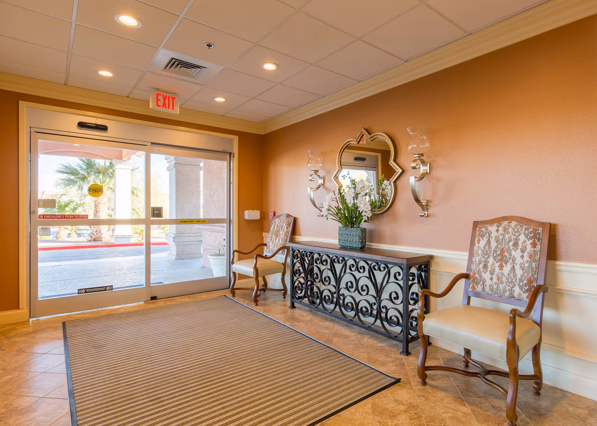 Entrance area of The Bridge at Paradise Valley featuring a glass automatic door with an exit sign above it. Inside, there are two upholstered wooden chairs on either side of a decorative black metal console table with a flower arrangement and a uniquely shaped mirror on the wall. The walls are painted a warm beige color and the floor is tiled with a large striped mat near the door.