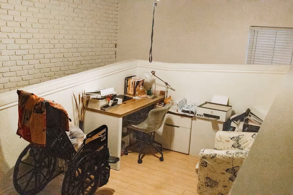 Small home office corner with a desk, office chair, printer, wheelchair, and a patterned armchair.