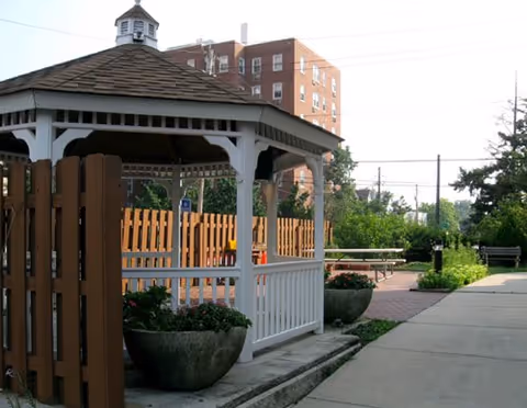 A white wooden gazebo and planters beside a paved walkway in a landscaped courtyard with a brick apartment building in the background.