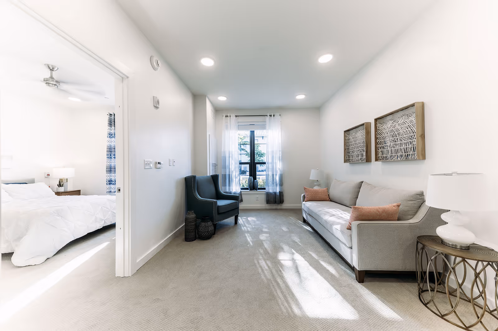 A bright living room with a gray sofa adorned with two orange pillows, a side table with a white lamp, and two framed artworks on the wall. Across from the sofa is a dark blue armchair with two decorative vases on the floor beside it. A window with sheer white curtains allows natural light to fill the room. To the left, an open doorway reveals a bedroom with a white bedspread, a nightstand with a lamp, and a ceiling fan.