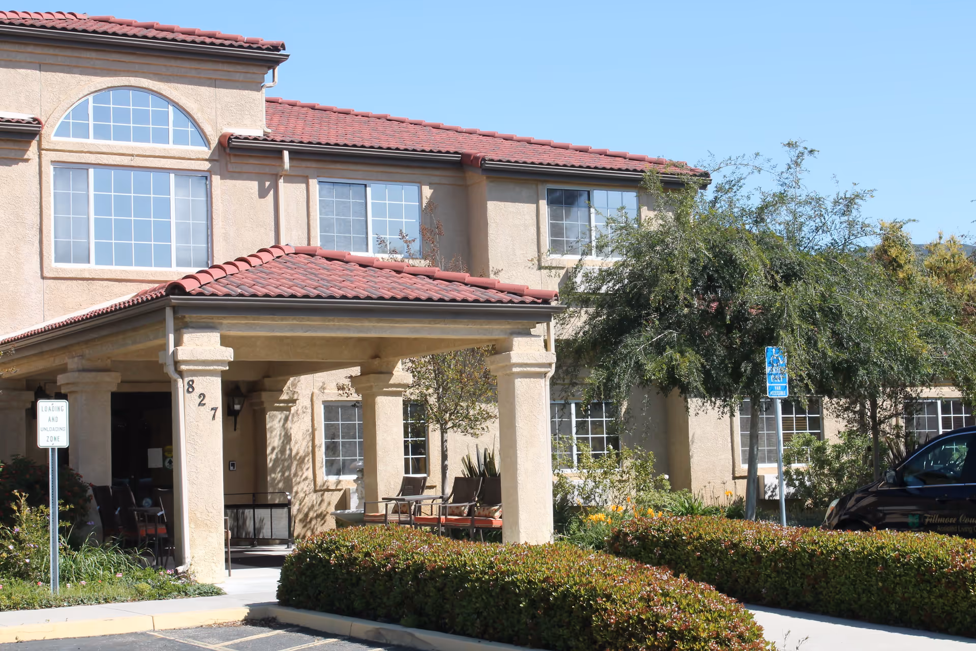 Exterior view of a two-story building with beige stucco walls and a red tiled roof. The entrance has a covered porch supported by columns, with the number 827 displayed on one column. There are several windows and some outdoor seating visible under the porch. Shrubs and trees surround the building, and a parking area is in front with a handicapped parking sign.