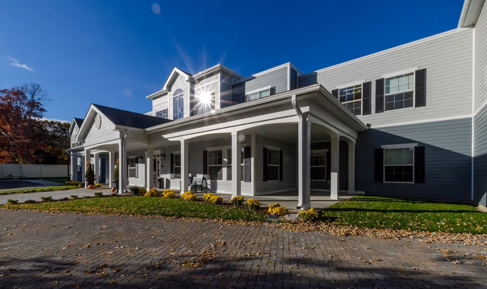 Exterior view of a senior living facility building with a covered porch, white columns, and multiple windows. The building is light gray with white trim and black shutters. There is a paved driveway and landscaped grass with yellow flowers in front. The sky is clear and blue.