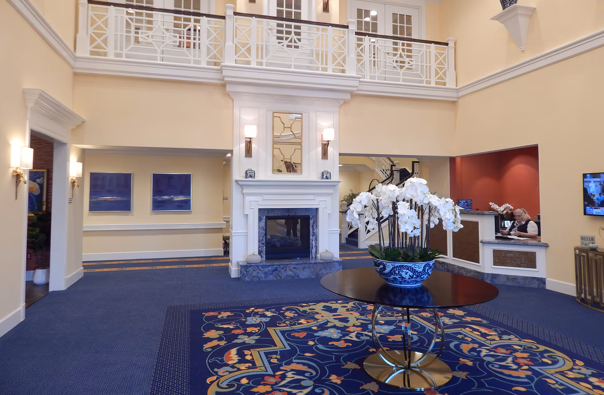 Spacious and elegant assisted living facility lobby with a blue patterned carpet, a round table with a large floral arrangement of white orchids in a blue and white vase, a white fireplace with a mirror above it, and a reception desk with a staff member working. The walls are painted light yellow, and there is a balcony with white railings overlooking the lobby.