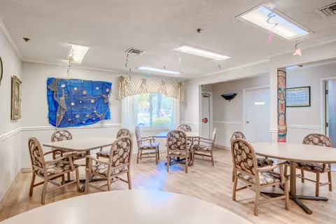 Bright communal dining/activity room with round tables and floral-upholstered chairs, wall decorations, and a window.