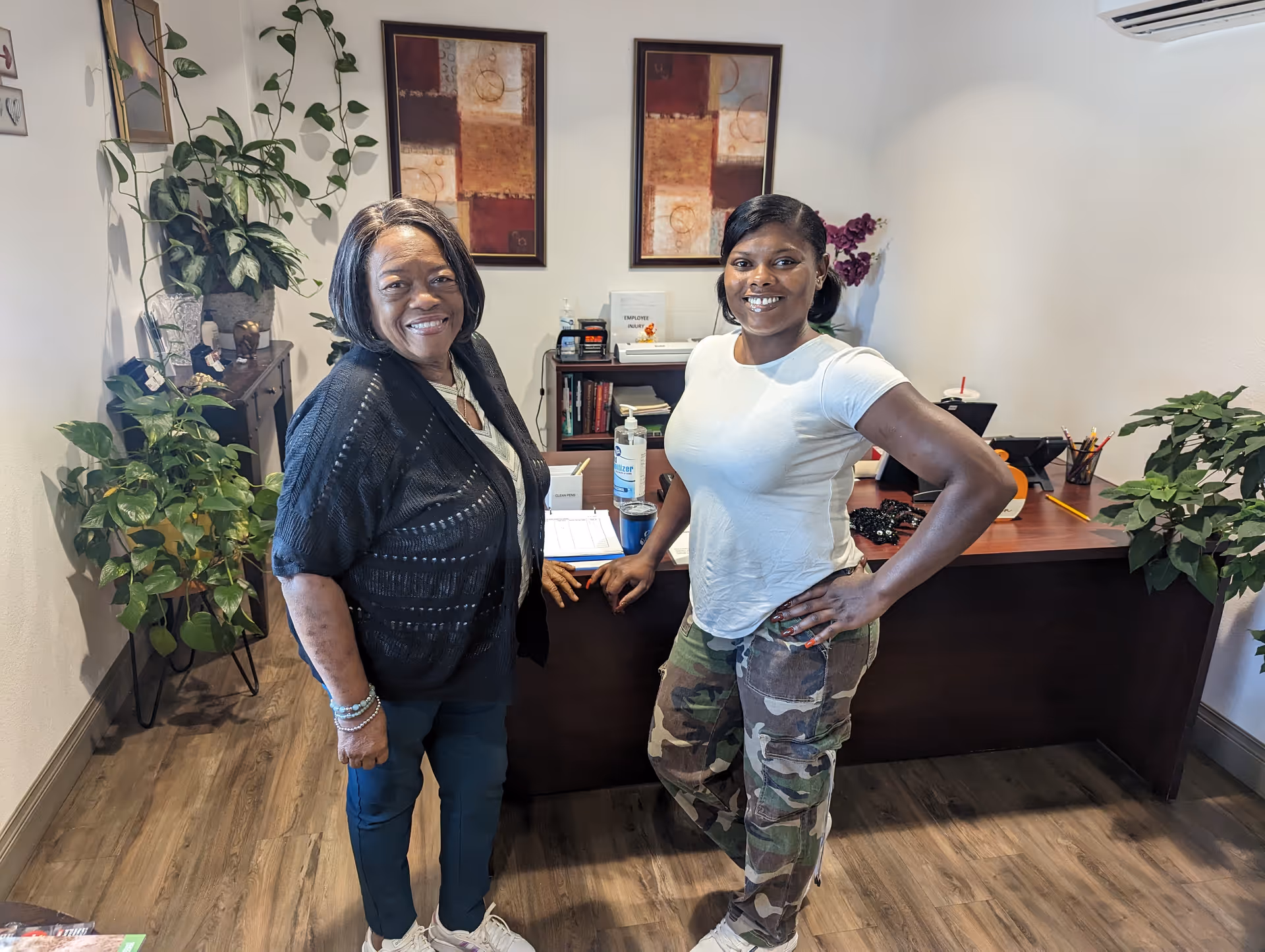 Two women smiling and standing in an office-like room with a wooden desk, plants, and framed artwork on the wall behind them.