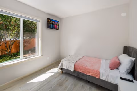 A small, bright bedroom with a single bed against the right wall. The bed has a gray upholstered headboard, white sheets, a pink blanket, and a pink pillow. A remote control rests on a white nightstand next to the bed. There is a large window on the left wall letting in natural light and showing a view of a garden with trees and a wooden fence. A small flat-screen TV is mounted high on the wall near the window. The room has light-colored wooden flooring and plain white walls.
