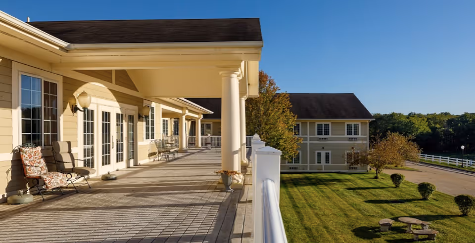 A spacious outdoor balcony or porch area of a senior living facility with several chairs and tables along the wall, overlooking a well-maintained lawn with trimmed bushes, trees, and a picnic table. The building is light yellow with white trim and large windows, under a clear blue sky.