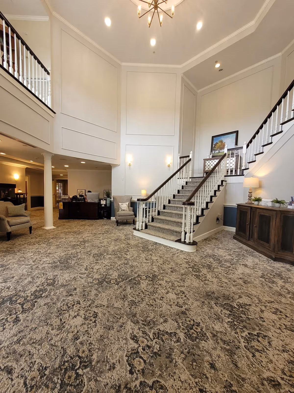 Spacious and elegant interior lobby area with a patterned carpet floor, a central staircase with white railings and dark wood handrails, beige walls with decorative molding, two armchairs, a wooden cabinet with a lamp and plants, and a chandelier hanging from the ceiling.
